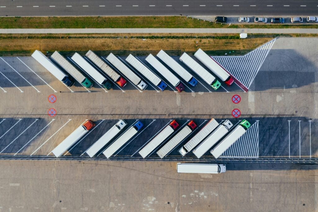 Aerial view of a parking lot filled with semi-trailer trucks parked in diagonal rows, adjacent to a road with cars and a sidewalk.