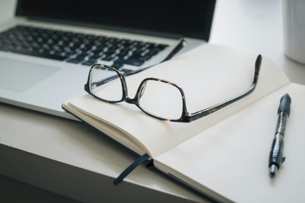 Eyeglasses resting on an open lined notebook beside a pen and a laptop on a white desk, suggesting a workspace or study setting.