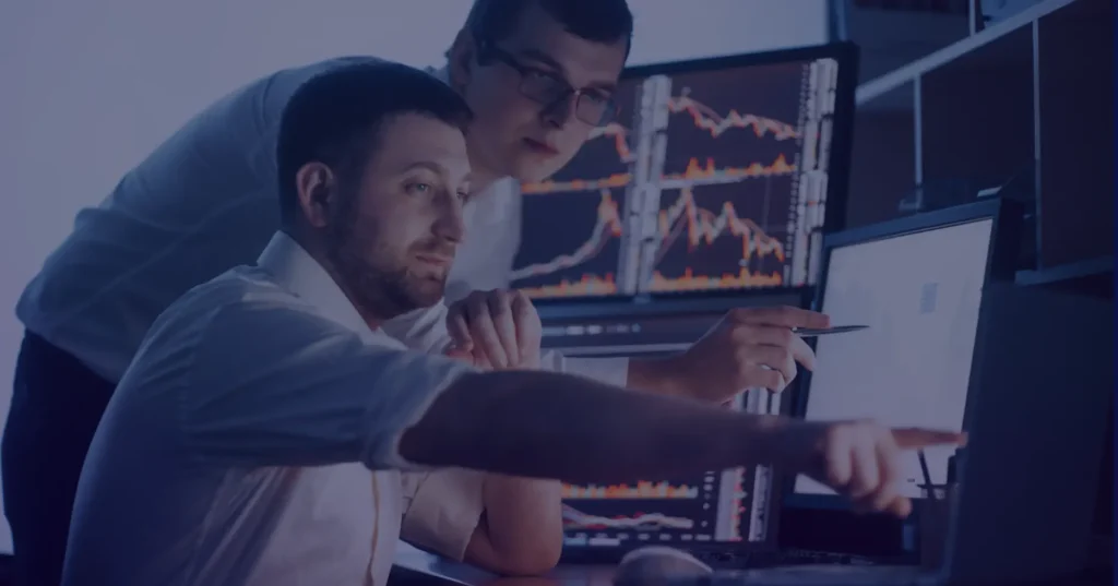 Team collaboration on financial data analysis, with one person pointing at a monitor displaying real-time stock market trends