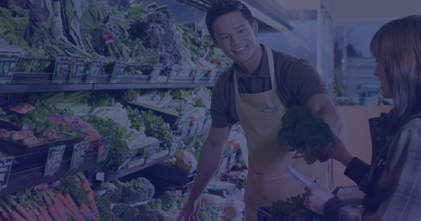 Smiling grocery store employee wearing an apron hands a head of broccoli to a customer in the produce section filled with fresh vegetables.