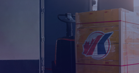Warehouse loading dock scene showing a pallet jack moving a shrink-wrapped pallet of cardboard boxes into the back of an open delivery truck. The pallet sits on a lift gate, and the boxes feature a logo combining a maple leaf and French flag colors, suggesting shipping or logistics operations.