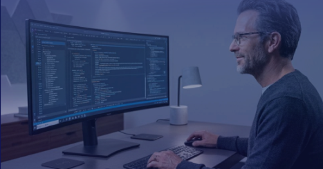 Man wearing glasses works at a desk on a desktop computer with a large monitor displaying code, in a softly lit office setting with a desk lamp and keyboard visible.