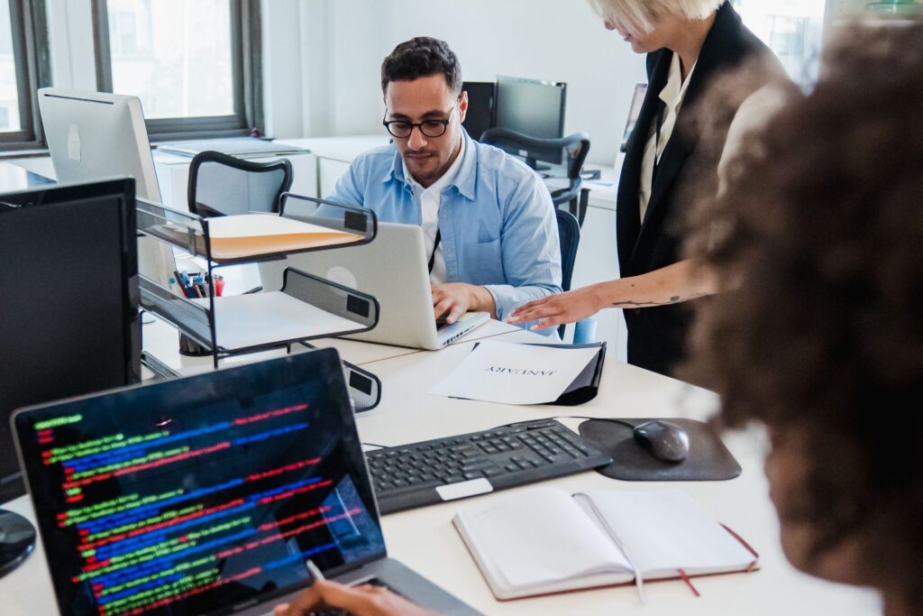 Two coworkers in an office working at laptops with colorful code on screens and documents on the desk.