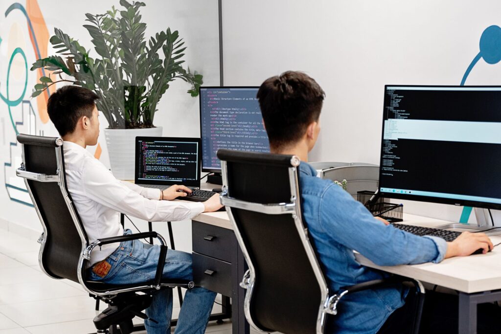 Two programmers seated at desks coding on multiple monitors in a bright office environment.
