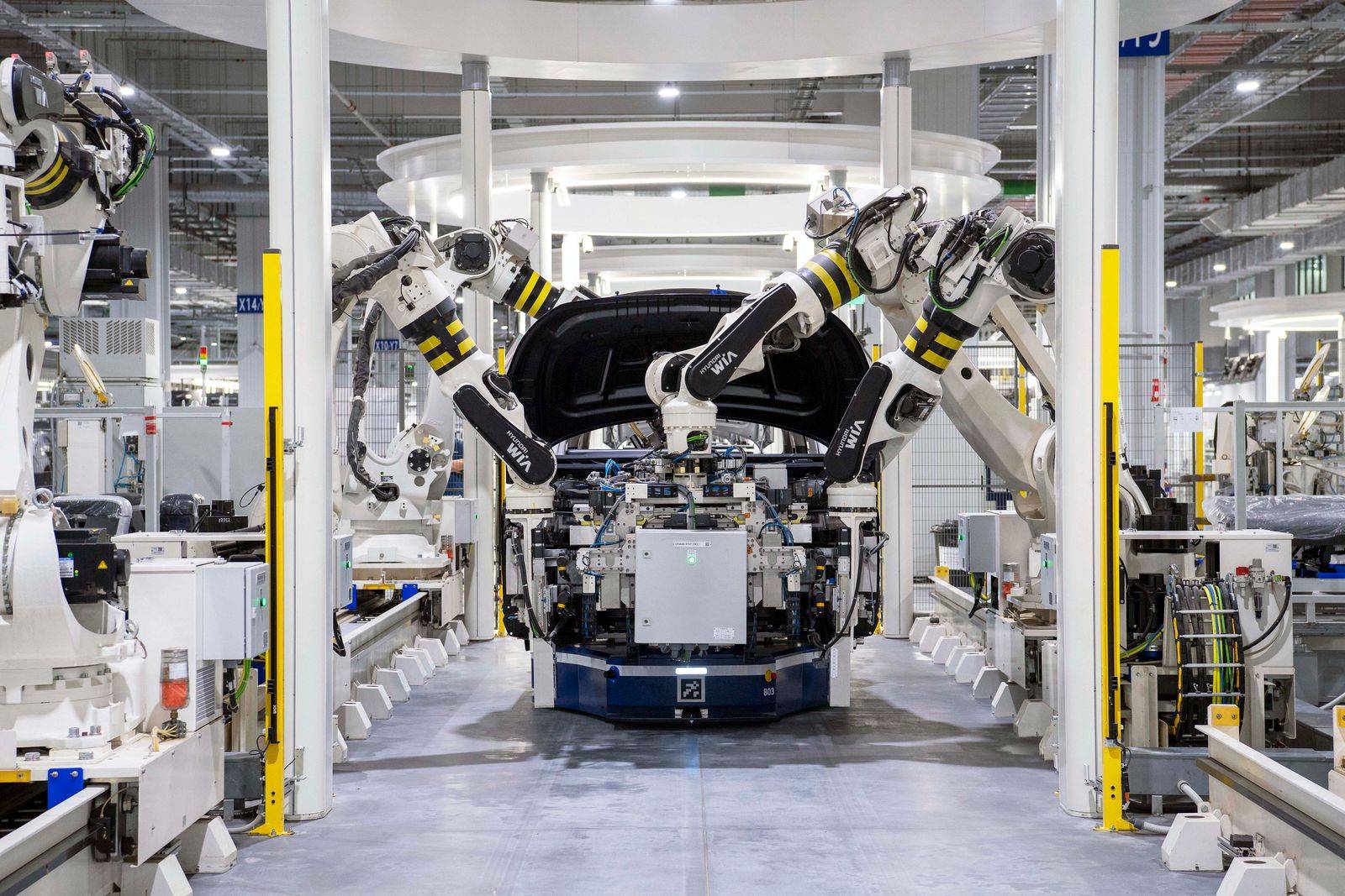 Six robotic arms with yellow and black stripes surround a car chassis on a high-tech production line, performing assembly tasks.