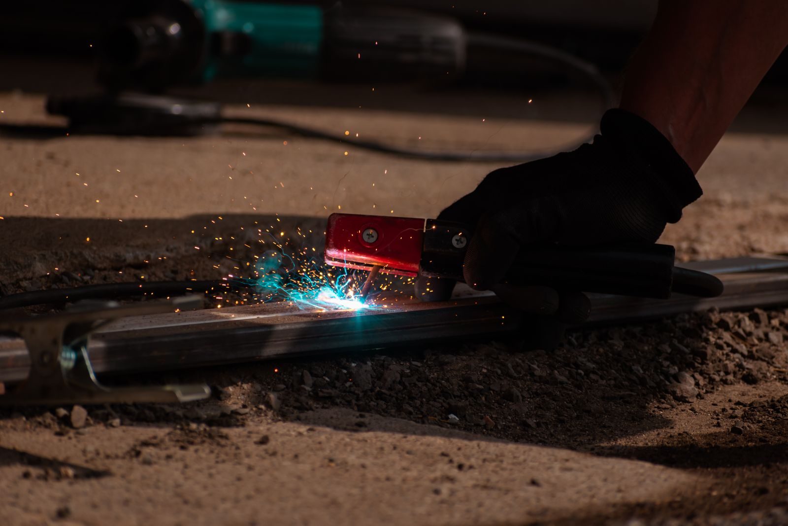 Worker in a glove uses a red handheld welding/grinding tool on a metal rail, sparks flying and a blue arc visible nearby.