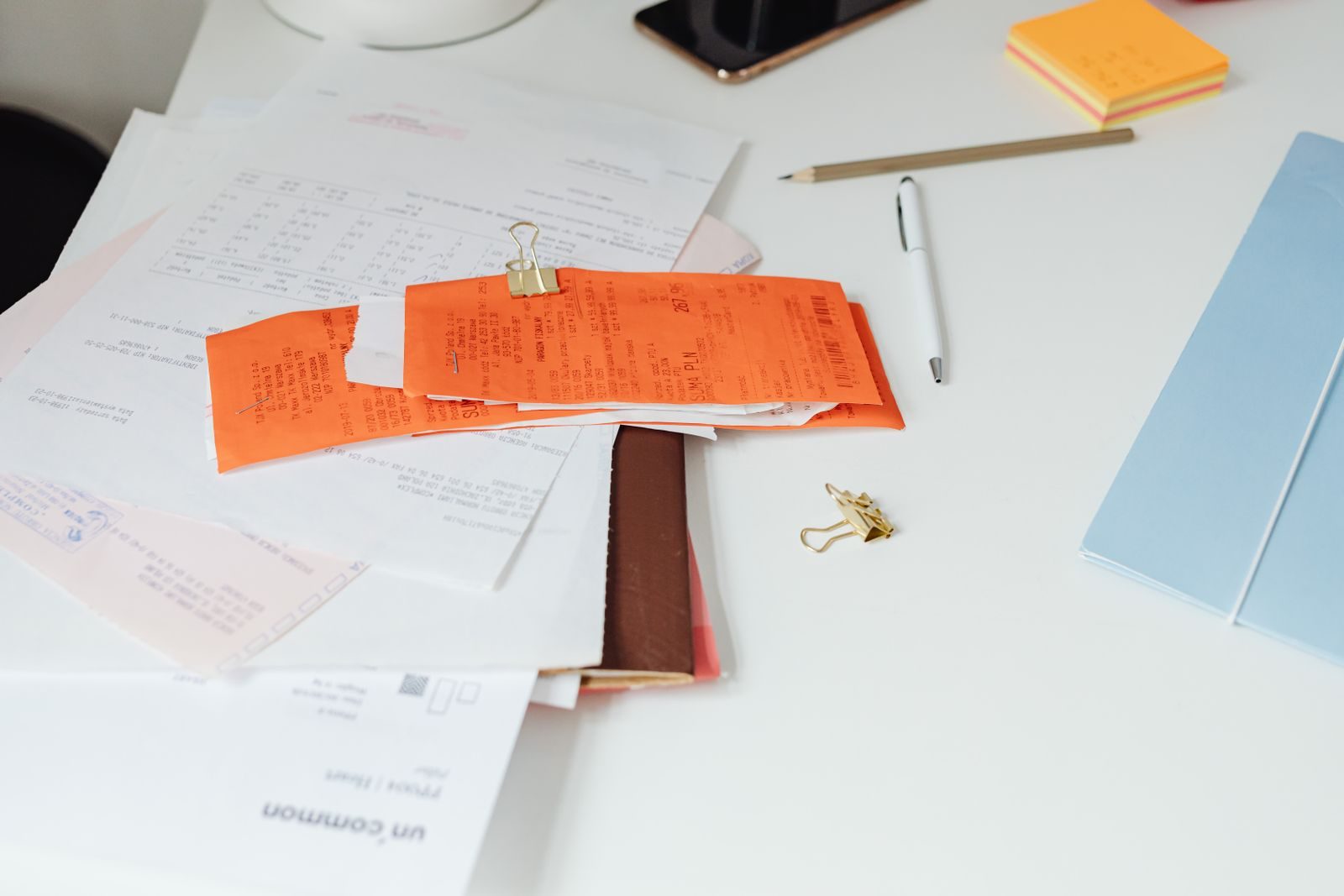 Desk with a pile of papers and orange receipts held by a gold binder clip, on a white desk with a pen, pencil, sticky notes, and a blue folder nearby