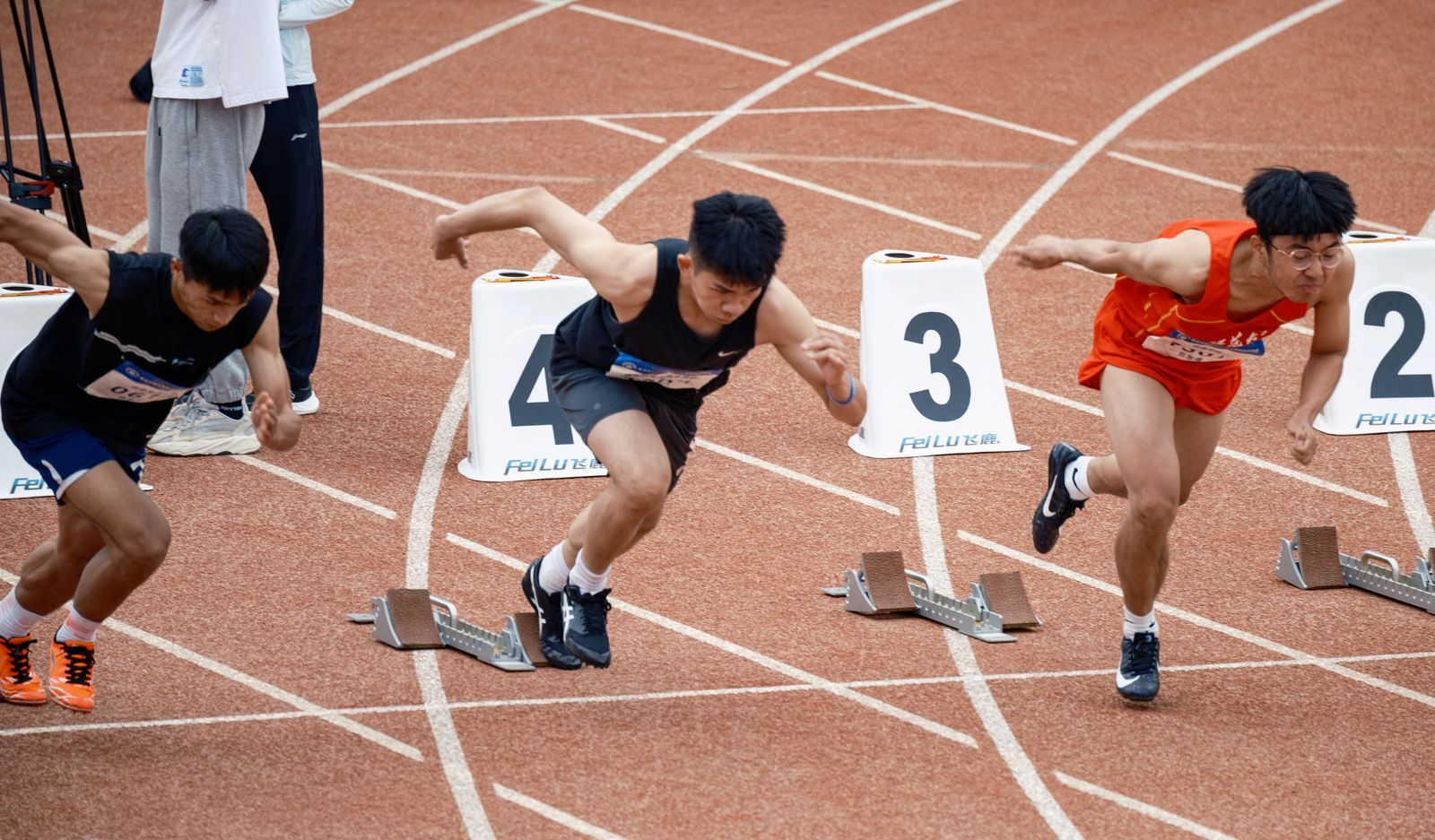 Three male sprinters start a race from starting blocks on a red track, wearing black and red uniforms, with blocks numbered 4, 3 and 2 nearby.