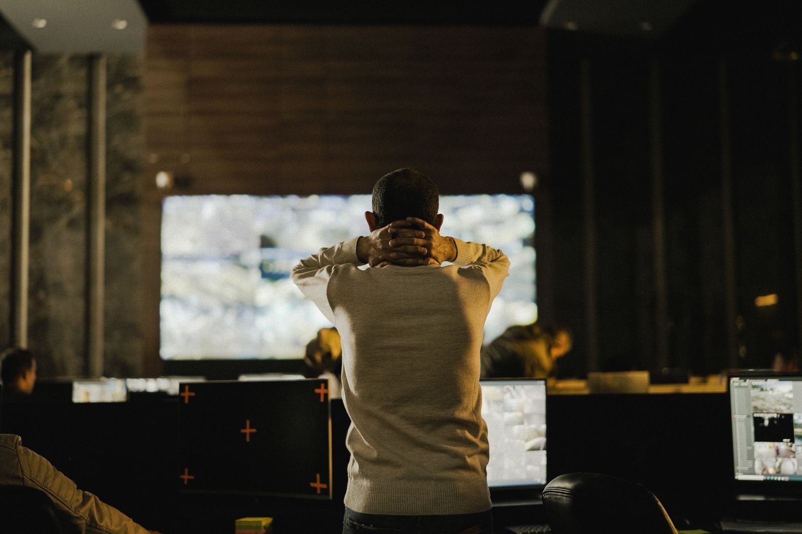 Man stands with hands clasped behind neck, facing a large illuminated screen in a dim control room or office.