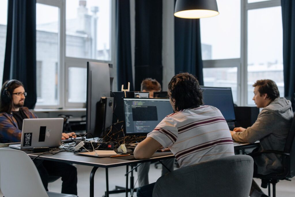 Software developers coding at shared desks with multiple monitors in a bright open office.