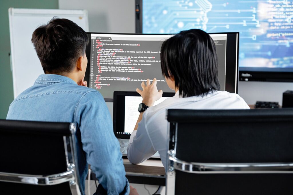 Two coworkers sit at a desk, reviewing code on a computer monitor in a modern office setting.
