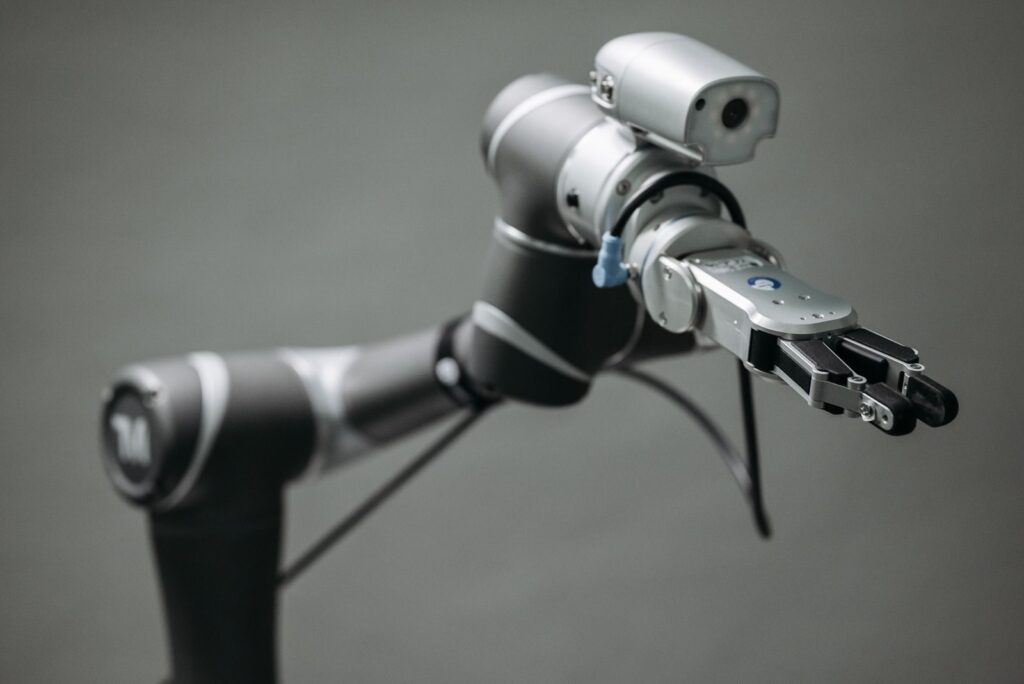 Silver robotic arm extends its clamp-style gripper toward the camera, blue tag visible near the joints, against a gray background.
