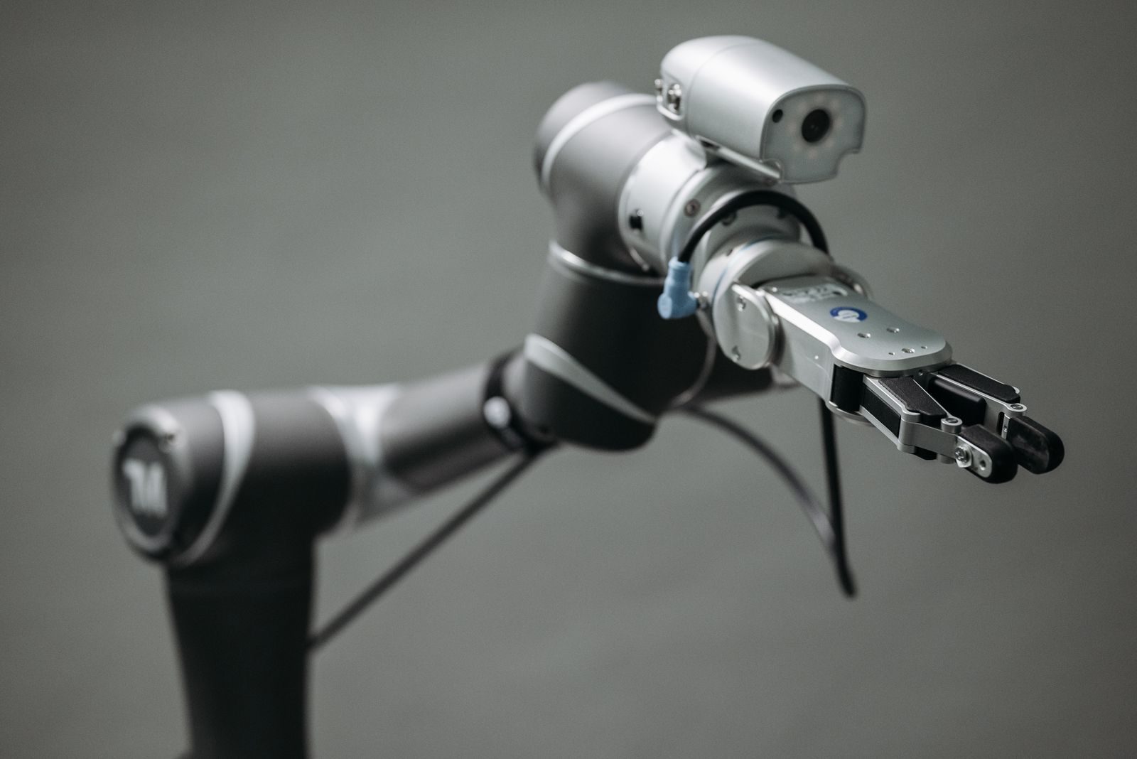 Silver robotic arm extends its clamp-style gripper toward the camera, blue tag visible near the joints, against a gray background.