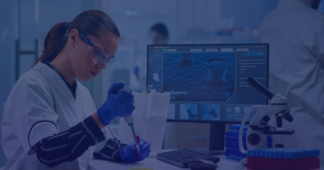 Scientist in a lab coat wearing safety goggles and gloves pipettes liquid at a workstation with a computer monitor displaying data.